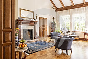 Blue armchairs and patterned carpet in front of wooden fireplace in sophisticated interior. Real photo