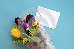 Blue background with composition made of blooming lilac and yellow tulips and white envelope.Gift