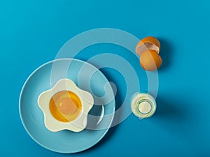 Blue background with egg in a flower shaped bowl and blue plate with broken egg shells and kitchen utensils