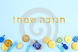 Blue background with multicolor dreidels, menora candles and chocolate coins with Happy Hanukkah wording in Hebrew