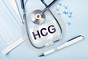 On a blue background, a stethoscope, a medical mask, pills, a pen and a white card with the text HCG Human Chorionic Gonadotrophin