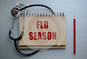 A blue background on which lies a notepad with the inscription FLU SEASON. Top view, floor plan. Stationery, stethoscope and plant