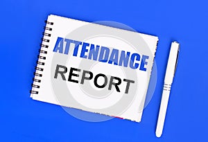 On a blue background, a white ballpoint pen and a white notepad with the text ATTENDANCE REPORT. View from above