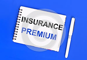 On a blue background, a white ballpoint pen and a white notepad with the text INSURANCE PREMIUM. View from above