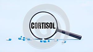 On a blue background, white and blue capsules with pills and a black magnifying glass with the text CORTISOL. Medical concept