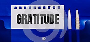 On a blue background, white pens and a sheet of paper with the text GRATITUDE