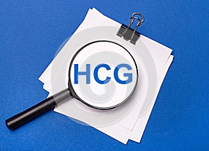 On a blue background, white sheets under a black paper clip and on them a magnifying glass with the text HCG Human Chorionic