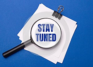 On a blue background, white sheets under a black paper clip and on them a magnifying glass with the text STAY TUNED
