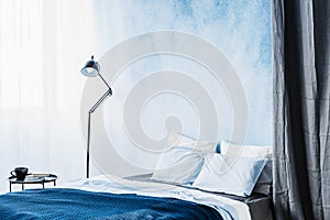 Blue bed between lamp and grey curtain in simple bedroom interior with table and ombre wall