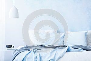 Blue bedsheets and pillows on bed next to a table with a cup under white lamp in simple bedroom interior