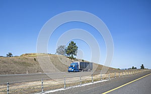 Blue big rig bonnet semi truck transporting cargo in tented black semi trailer running on the summer road with dry grass and along