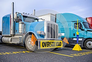 Blue big rig classic semi truck with oversize load sign on the bumper standing on the reserved spot on the truck stop beside