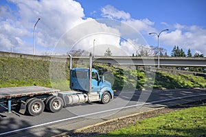 Blue big rig day cab semi truck with vertical exhaust pipes transporting empty flat bed semi trailer moving on the one line road