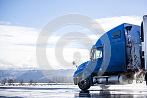 Blue big rig long haul semi truck with reefer semi trailer running on winter wet road with melting snow and water dust