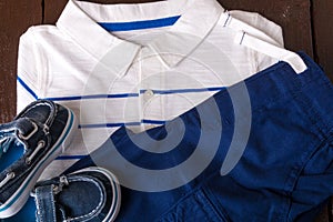 Blue boat shoes near shorts and white striped polo on brown wooden background. Boy outfit. Top view.