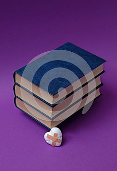 Blue books, hardcover, stacked horizontally, one on top of the other, a broken little white heart with patches, on a purple