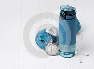 Blue bottle of water, towels, headphones and stopwatch. Sports equipment on white wooden background