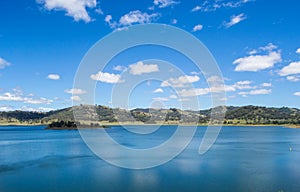 Blue calm deep water lake with small island surrounded by hills with trees under a blue sky with small soft white clouds.