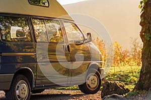 A blue Camper Van sits at sunset against a bright green meadow with lush orange sunlight streaming in