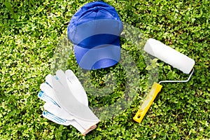 Blue cap, paint roller and white working gloves on green grass.