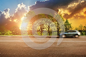 A blue car driving fast on the countryside road against sky with sunset