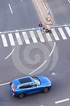 Blue car heading to pedestrian crossing with unrecognizable young woman pushing baby carriage, safety of self-driving technology