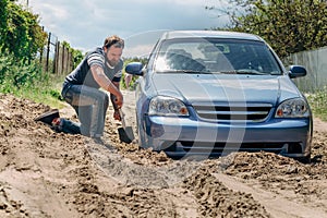 A blue car stuck in the sand.. A strong man with a beard digs a shovel of the wheel of the car.