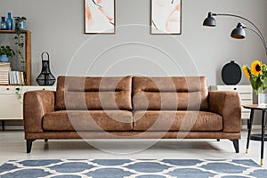 Blue carpet in front of brown leather sofa in grey living room interior with posters. Real photo