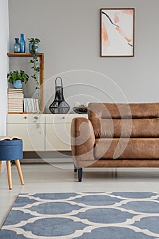 Blue carpet and stool in grey living room interior with plants and poster above couch. Real photo