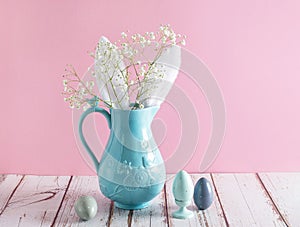 Blue ceramic jar with rabbit ears and bouquet of white small flowers of Gypsophila paniculata on pink background. Nearby wooden