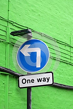 Blue circular road sign with a left turn arrow and notice saying one way against a green brick wall