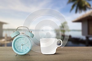 Blue classic alarm clock and hot white coffee cup with steam on vintage wooden desk on blurred beach background