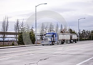 Blue classic big rig American semi truck running on wide road transporting two covered bulk semi trailer with commercial cargo