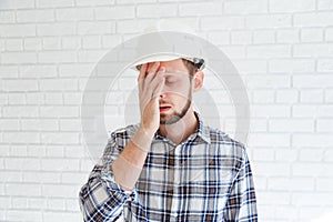 A blue collar worker wearing a hardhat is covering his face with his hand
