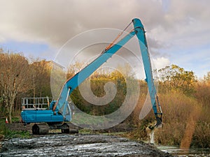 Blue color excavator working on deepening and cleaning a small river, Sunny day, Cloudy sky