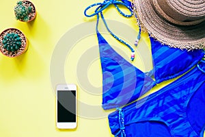 Blue color swimwear with smartphone, cactus and hat flat lay on yellow background, Summer vacation