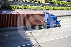 Blue comfort long haul big rig semi truck with long container driving on the divided highway road running under bridge