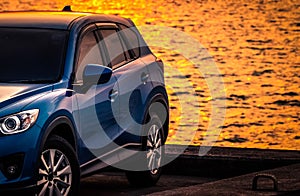 Blue compact SUV car with sport and modern design parked on concrete road by sea at sunset. Environmentally friendly technology.