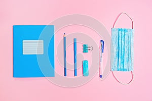 Blue copybook, protective mask and office supplies on pink background. Flat lay. Back to school banner. New normals of