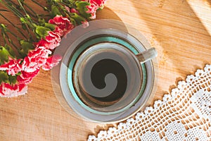 Blue cup of coffee, doily, pink carnation flowers on wooden table illuminated by sun light