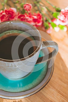 Blue cup of coffee, pink carnation flowers on wooden table illuminated by sun light
