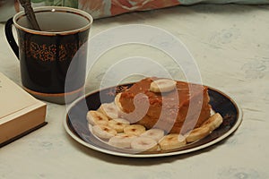 A blue cup of hot tea, a piece of shortbread cake with condensed milk and bananas, cut rings on a saucer and an open book with whi