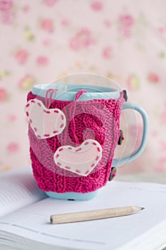 Blue cup in pink sweater with felt hearts standing on notebook
