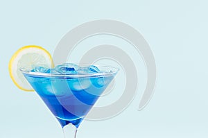 Blue curacao cocktail with ice cubes, lemon slice in luxury martini glass on pastel mint color wall, closeup, edge, details.