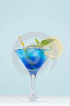 Blue curacao cocktail with ice cubes, lemon slice, yellow straw, green mint in luxury martini glass on white wood board and green.