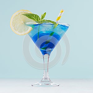 Blue curacao cocktail with ice cubes, lemon slice, yellow straw, green mint in luxury martini glass on white wood board and pastel