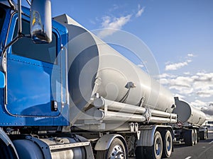 Blue day cab big rig semi truck transporting liquid cargo in two tank semi trailers moving on the wide road