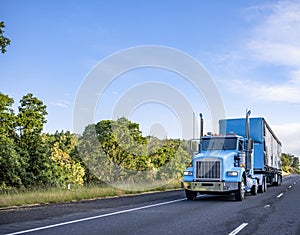 Blue day cab classic big rig semi truck transporting cargo in blue semi trailer moving on the straight highway at sunny day