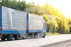 Blue delivery van and white truck driving on the asphalt road between line of trees in autumn landscape