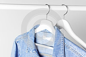 Blue denim jacket on white wooden coat hanger on a rod against light gray wall flat lay copy space. Denim, fashionable jacket,